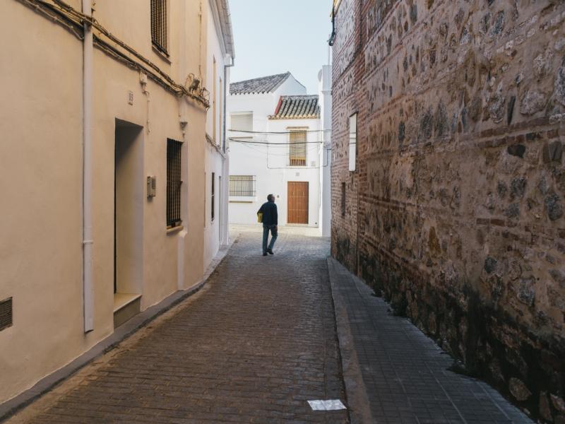 Calle del municipio, casa de paredes encaladas a un lado y muro de piedra de edificio histórico al otro, pavimento adoquinado, persona caminando