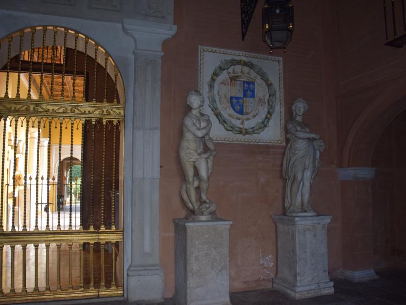 Imagen de una puerta con su reja dorada y dos figuras de mármol a la entrada además del escudo  heráldico